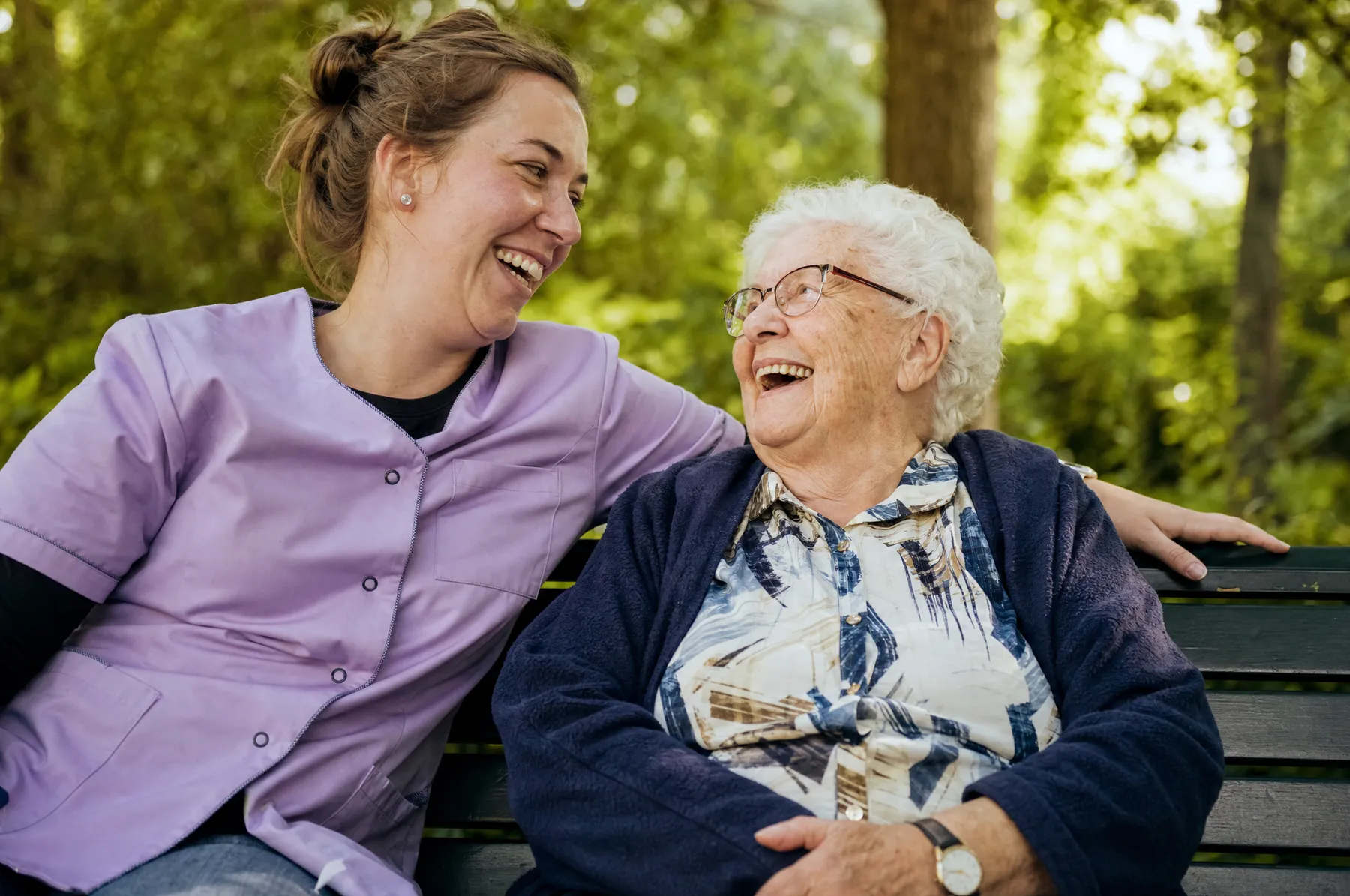 Zorgverlener en oudere vrouw lachen samen op een bankje in de tuin